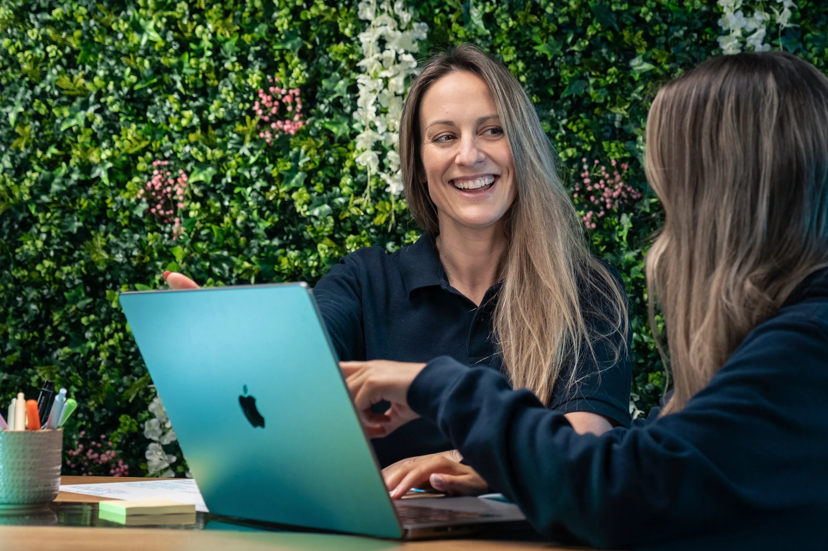 Rachel Fabrizio in discussion with a colleague at the DotPerformance office in Douglas, Isle of Man, working on an Apple MacBook in front of a living plant wall